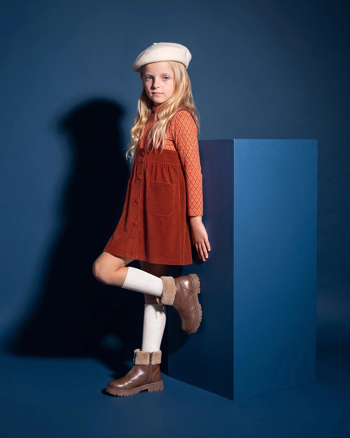 Puvu Pior® luxury girls' fashion against a deep blue studio backdrop, featuring a young blonde girl in a Rust Red corduroy pinafore jumper dress over an geometric-patterned long-sleeve top, styled with a cream beret, white knee-high socks, and brown shearling-lined boots.