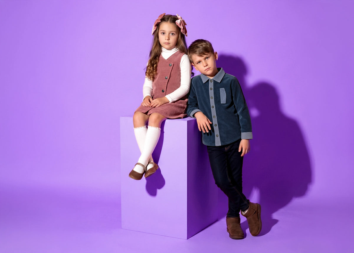 Puvu Pior® luxury children's collection against a vibrant purple backdrop, featuring a young girl sitting on a block in a dusty rose corduroy overall pinafore dress with a white turtleneck, knee-high socks, and velvet hair bows, next to a young boy leaning in a dark slate-blue corduroy button-up shirt with contrast trims and dark denim jeans.