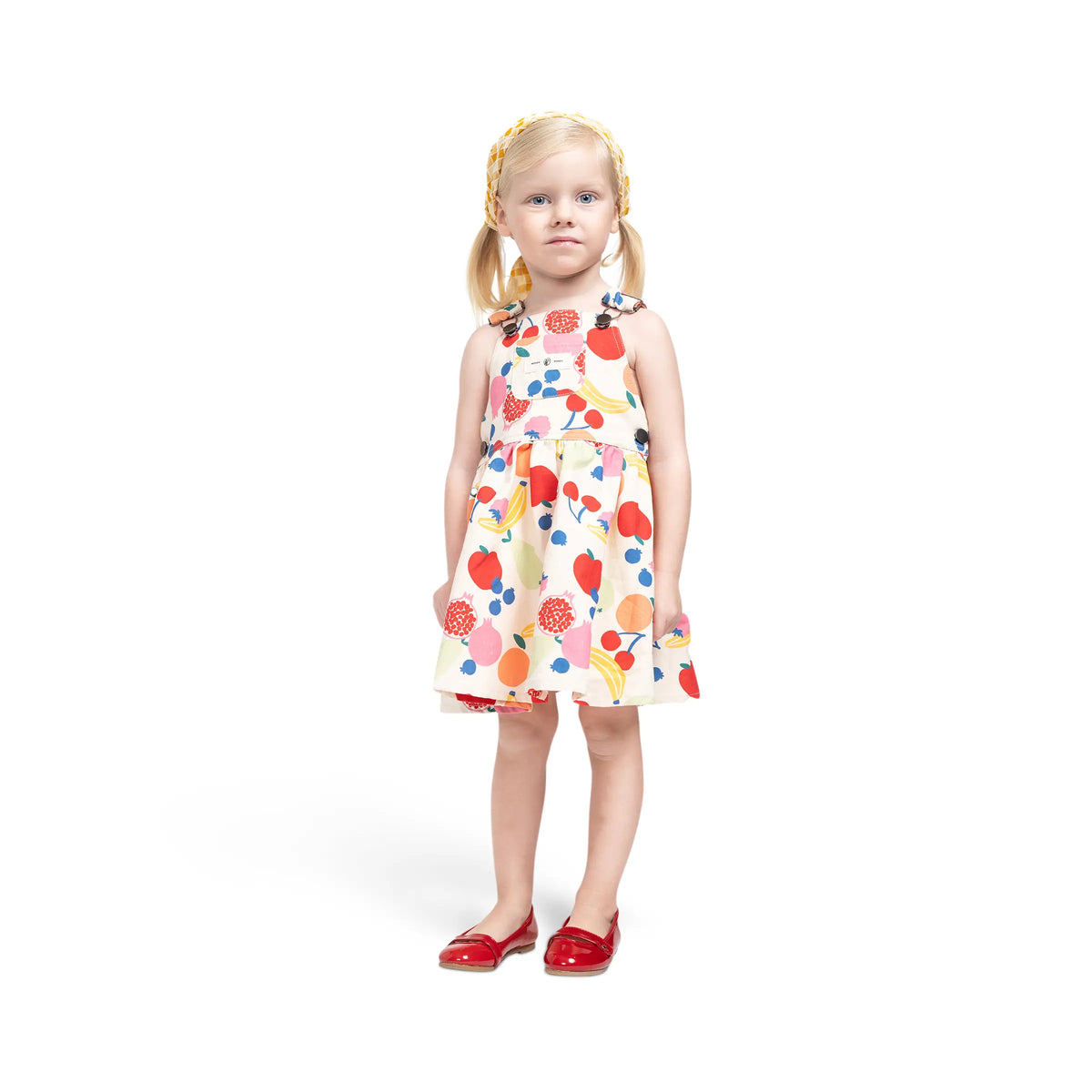 Girl modeling Puvu Pior® colorful fruit print pinafore, styled with a yellow headscarf and red shoes, showing fit.